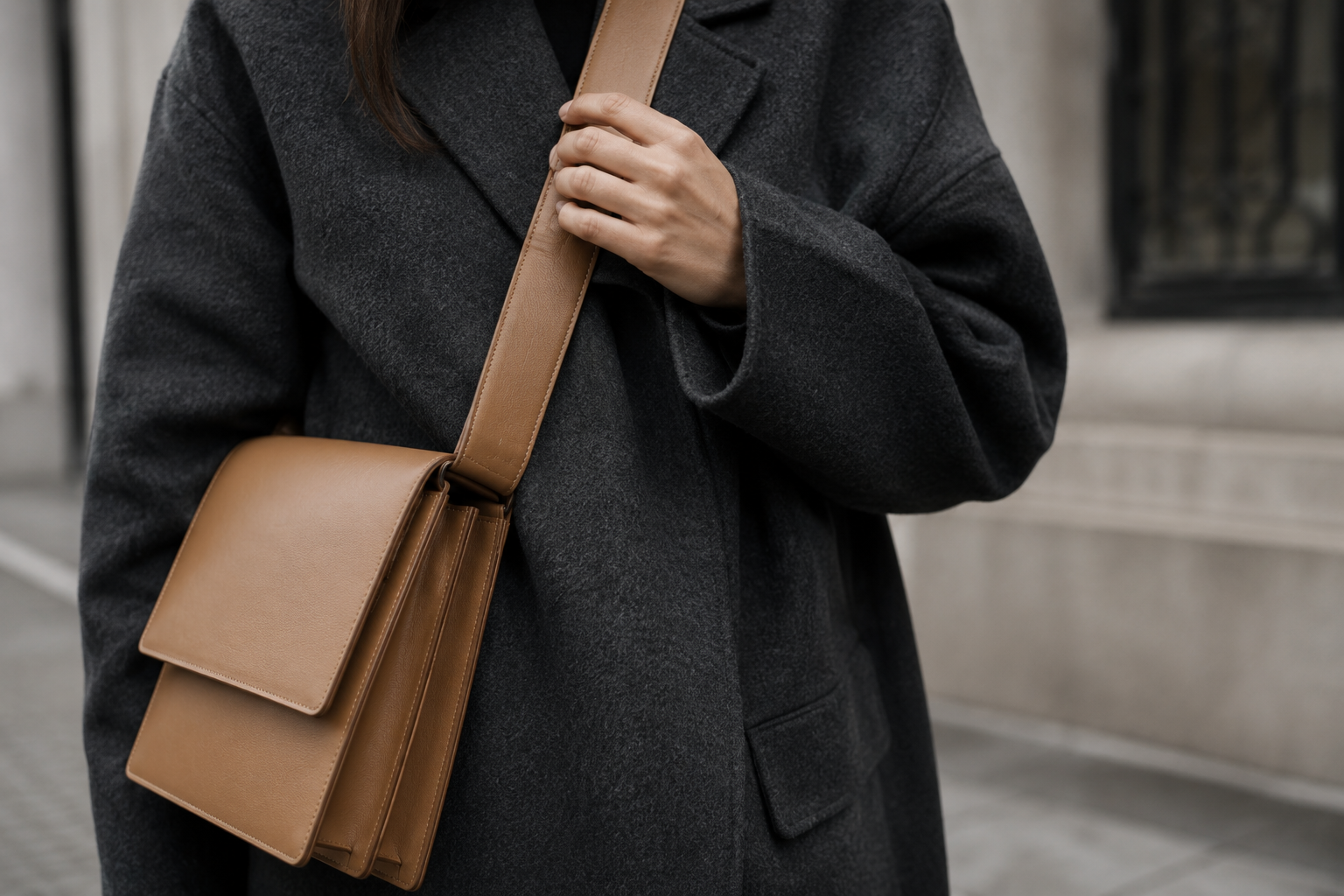 Close-up of a hand adjusting the wide leather strap of a tan structured crossbody bag worn over a charcoal wool coat outdoors