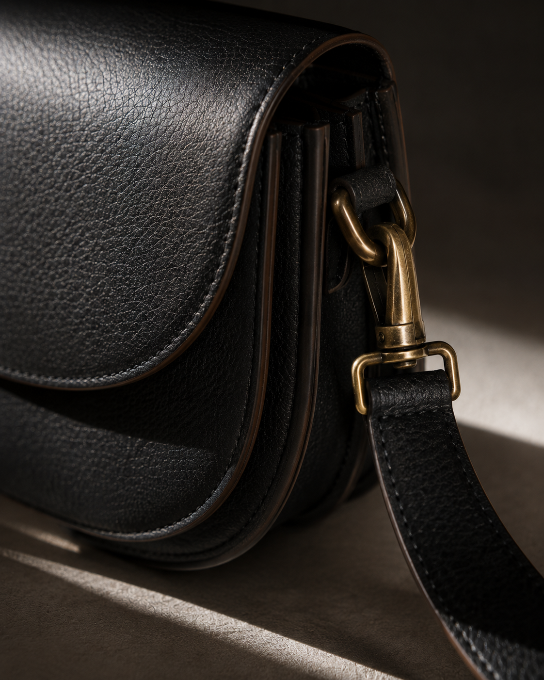 A tight detail shot showing grained black calfskin leather, polished brass hardware, and hand-painted edge finishing on a small crossbody bag