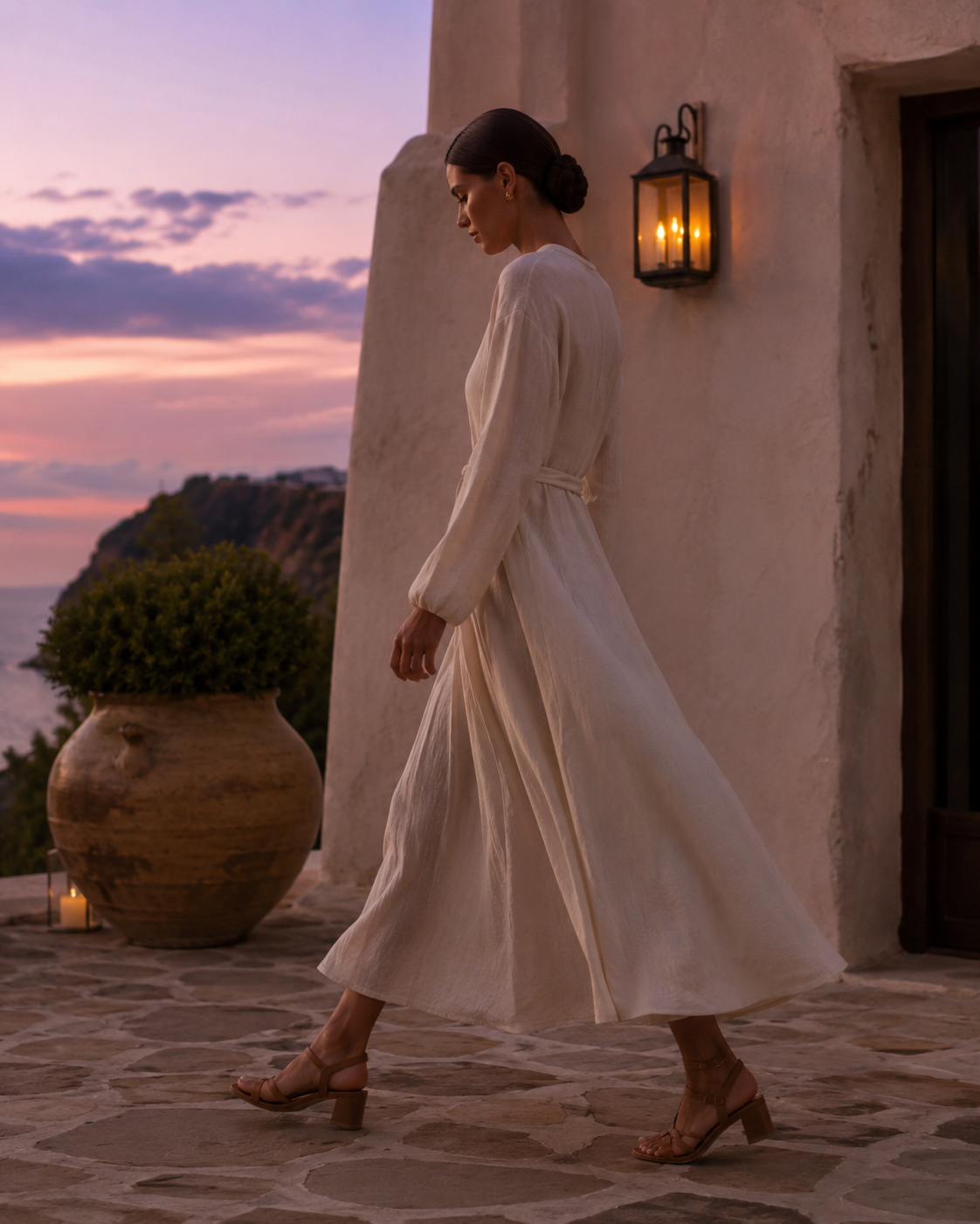 Woman in a cream linen maxi dress walking across a stone hotel terrace at dusk wearing tan leather mid heel sandals against a pink and lavender sky