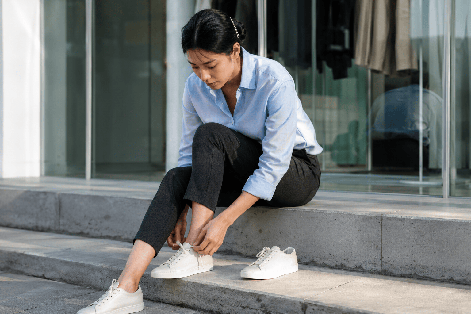 Vietnamese-French woman lacing white leather designer sneakers on a polished concrete step in front of a glass storefront