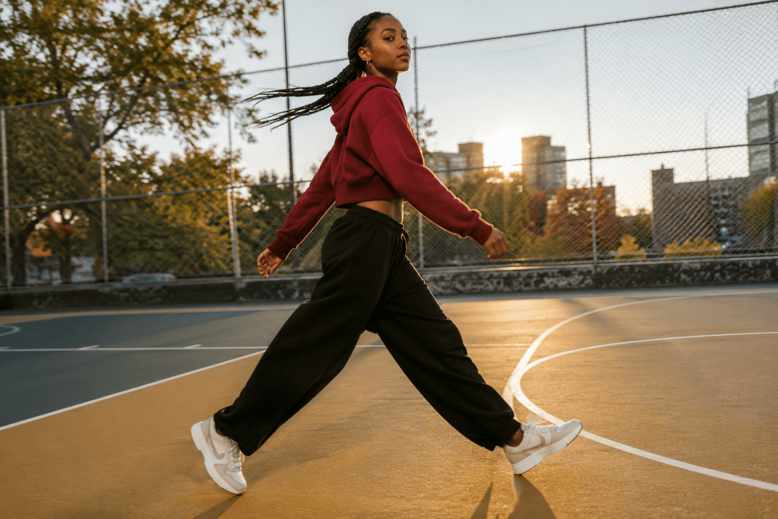 Caribbean teenage girl in maroon hoodie stepping onto a public basketball court at golden hour in white-and-tan leather sneakers