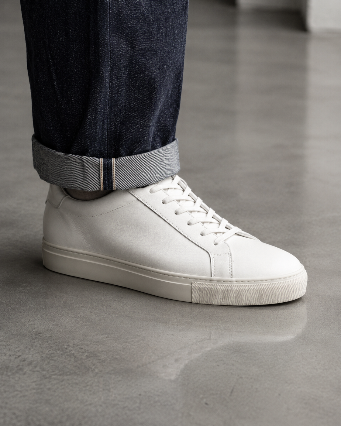 White leather low top sneaker styled with a raw denim cuff on a polished concrete studio floor