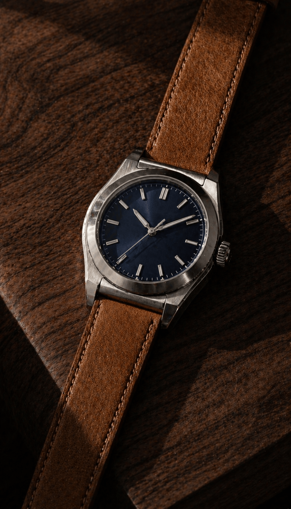 Close-up of a brushed stainless steel watch with a sunburst silver dial, applied indices, and a steel three-link bracelet resting on a worn oak desk